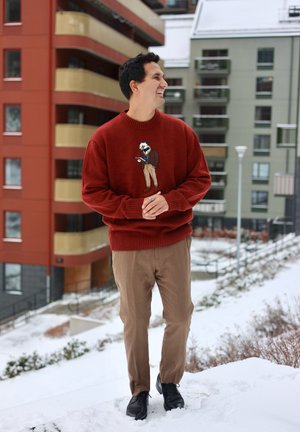 Hombre con suéter rojo con gráfico de animal y pantalones marrones sonríe al aire libre sobre suelo nevado cerca de edificios residenciales.