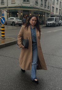 Manteau beige par-dessus une chemise en denim et un jeans évasé. Sac à main noir, assorti à des chaussures noires. Cadre urbain avec un café en arrière-plan.