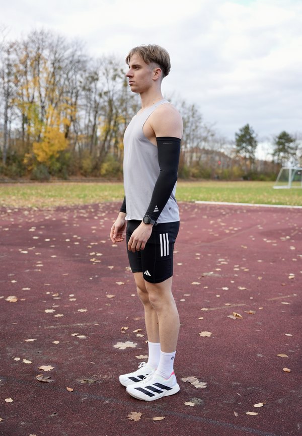 Juodas sportinis rankovės apatinis drabužis, pilkas marškinėlių liemenė, juodi šortai su baltais dryžiais ir balti sportbačiai su juodais akcentais. Lauko bėgimo takas.