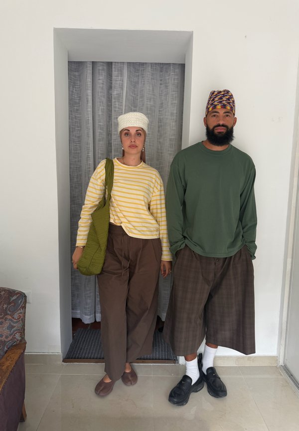 Mujer con camisa de rayas amarilla y pantalones marrones con gorro de punto blanco y bolso verde está junto a un hombre con camisa verde, pantalones de cuadros y gorro con estampado.