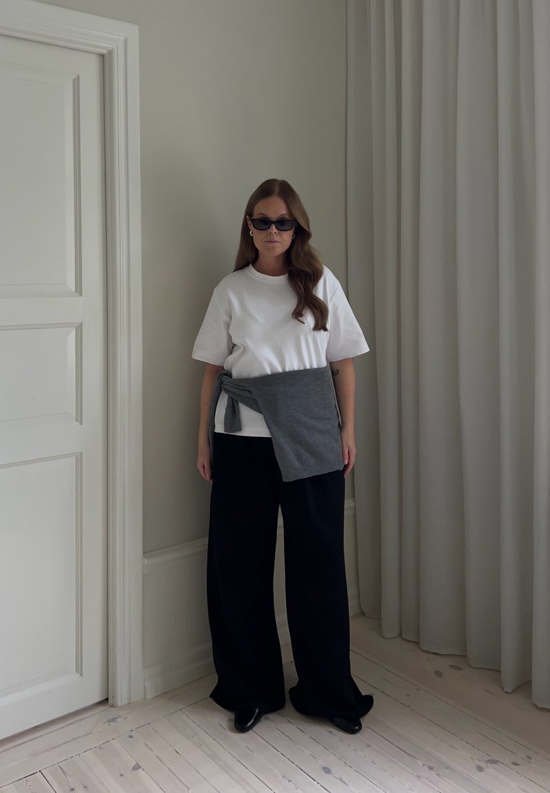 Femme portant un t-shirt blanc, un pantalon noir ample, un pull gris noué à la taille et des lunettes de soleil foncées, debout à l'intérieur près d'une porte fermée et de rideaux.
