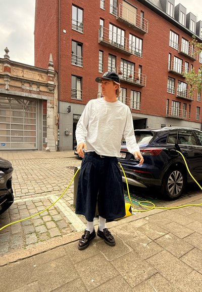Culottes de mezclilla, camisa blanca de manga larga, gorra negra, zapatos negros y calcetines blancos. Entorno urbano con un coche estacionado y un cable de carga amarillo.