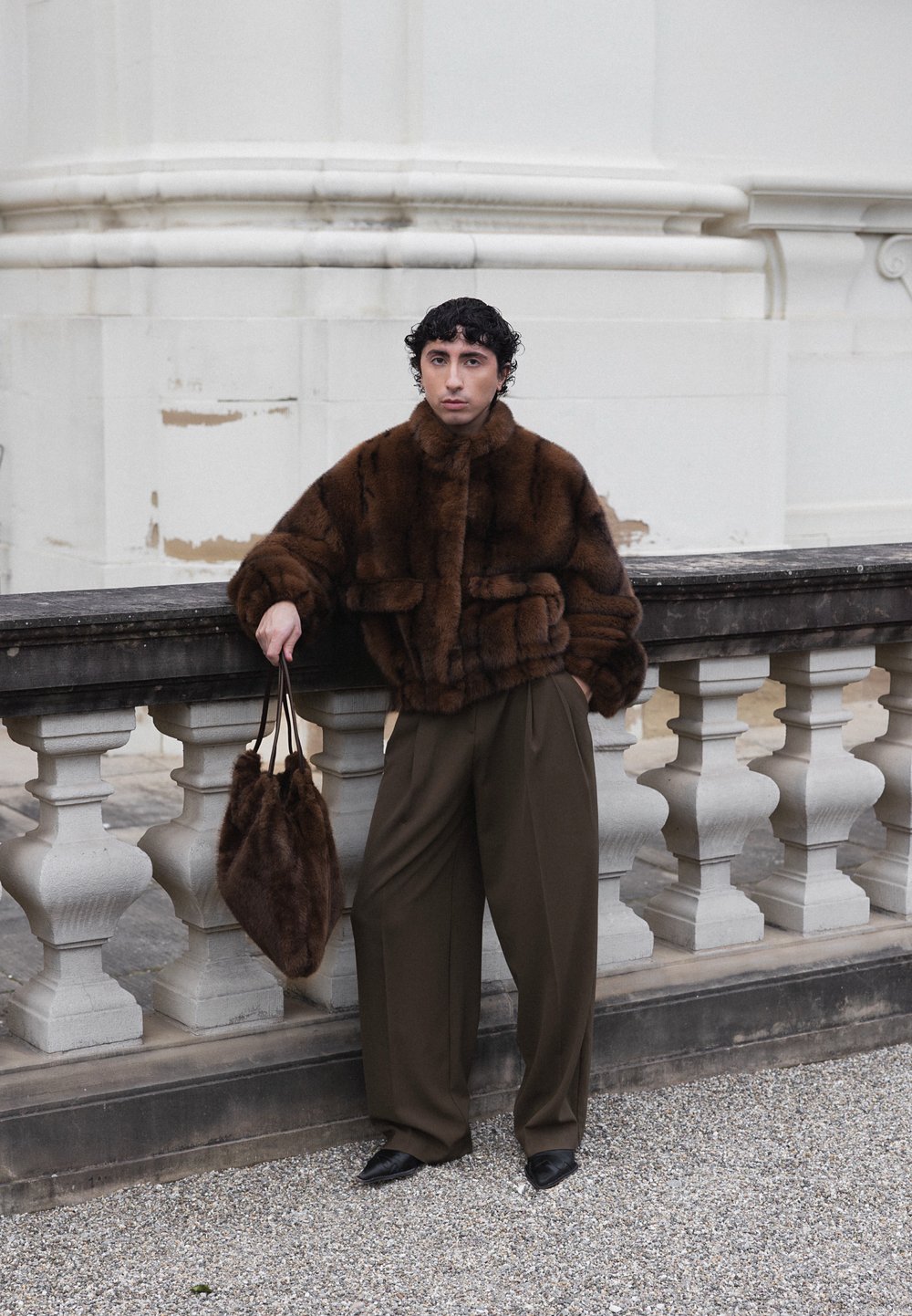 Person med kort, krøllet hår, læner sig op ad en stenskinne og har en mørkebrun pelsjakke, løse brune bukser, sorte sko og holder en matchende pels taske.