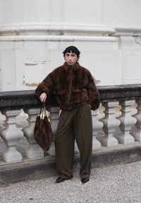 Person med kort, krøllet hår, læner sig op ad en stenskinne og har en mørkebrun pelsjakke, løse brune bukser, sorte sko og holder en matchende pels taske.