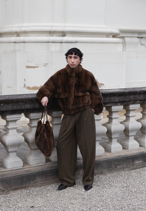 Person mit kurzen lockigen Haaren lehnt an einem steinernen Geländer, trägt eine dunkelbraune Pelzjacke, lockere braune Hose, schwarze Schuhe und hält eine passende Pelztasche.