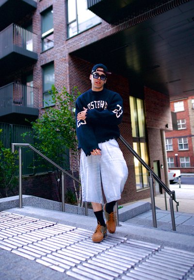 Suéter negro con letras blancas, pantalones cortos de mezclilla crema desgastados, zapatos marrones y gafas de sol; de pie en unos escalones exteriores con barandilla de metal.