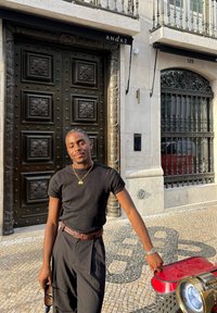 Maglietta nera a costine, pantaloni neri e cintura marrone. In piedi accanto a un veicolo rosso, con una porta nera ornata e una decorazione in pietra sullo sfondo.