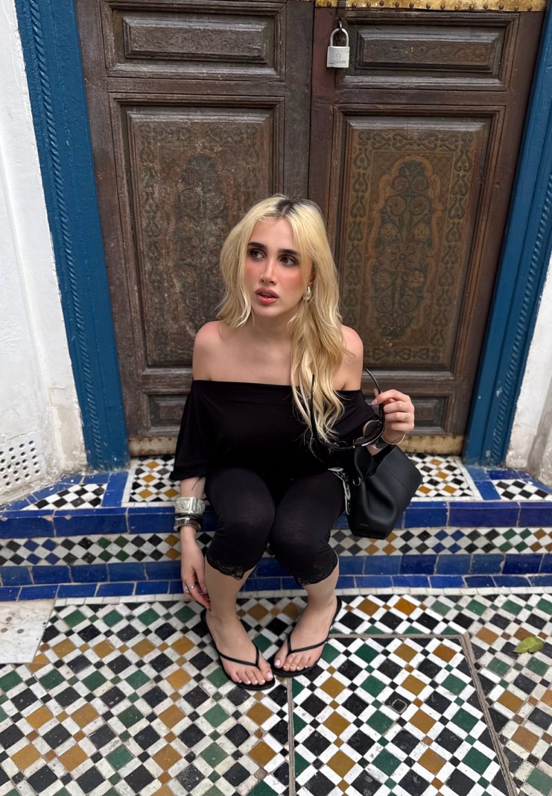 Blonde woman in black off-shoulder top and leggings sits on tiled steps in front of a wooden door, holding sunglasses and a black bag.