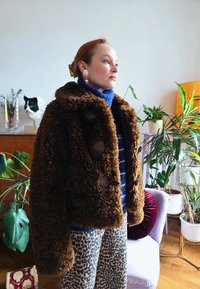 Femme portant une veste en fourrure marron, un col roulé bleu et un pantalon à imprimé léopard, debout dans une pièce avec des plantes et des objets décoratifs.