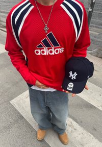 Osoba v červenej dlhorukáve Adidas tričku, vo voľných modrých džínsach, s béžovými topánkami, drží tmavomodrú čiapku New York Yankees so samolepkou značky 47.