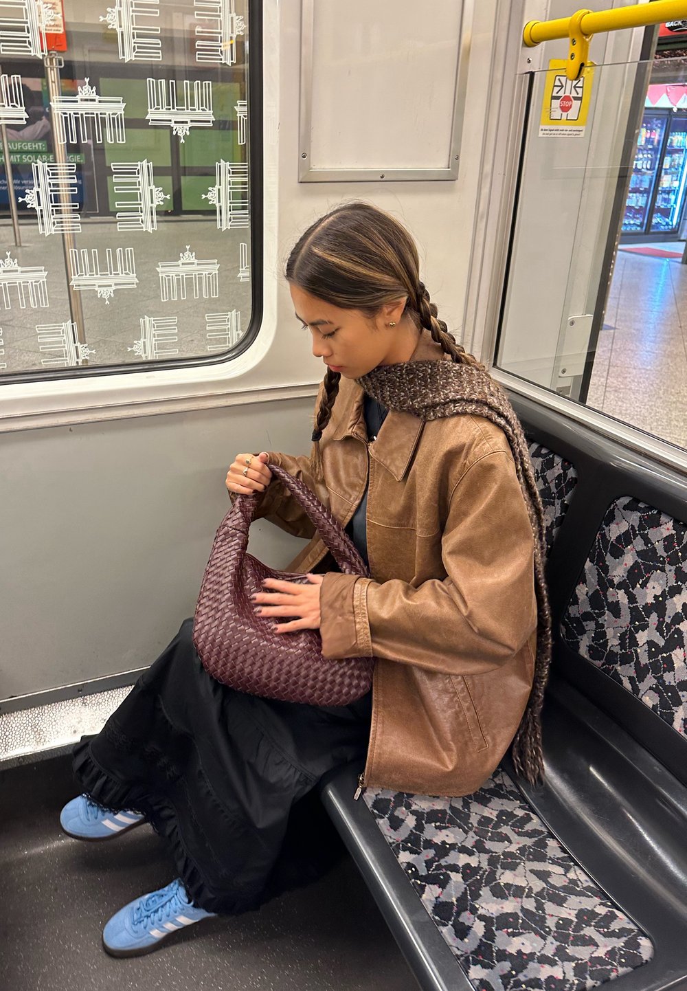 Bruine leren jas, zwarte gelaagde rok en blauwe sneakers. Houdt een geweven kastanjebruine tas vast, zittend op een gestreepte metrozit.