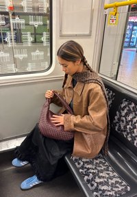 Bruine leren jas, zwarte gelaagde rok en blauwe sneakers. Houdt een geweven kastanjebruine tas vast, zittend op een gestreepte metrozit.