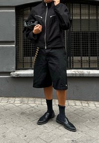 Osoba u crnoj jakni s patentnim zatvaračem, crnim šortsevima s zlatnim lancem, crnim čarapama i crnim lofericama stoji na trotuaru.