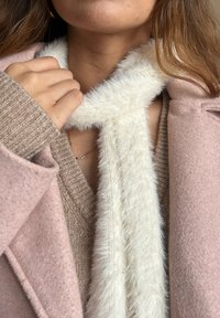Weißer, flauschiger Schal, der um den Hals gelegt ist, kombiniert mit einem beigen Strickpullover und einem hellrosa Mantel mit V-Ausschnitt.