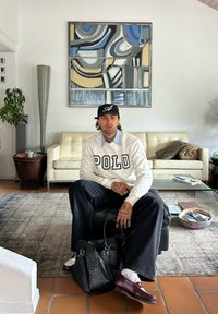 Mann sitzt auf schwarzem Ottoman im Wohnzimmer, trägt weißes Polo-Sweatshirt, schwarze Mütze und burgunderfarbene Schuhe, hält eine geflochtene schwarze Tasche.