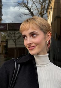 Junge Person mit kurzen blonden Haaren, die einen weißen Rollkragenpullover und einen schwarzen Mantel trägt, lächelt und schaut im Herbst draußen zur Seite.