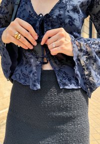 Navy bloes met bloemenpatroon van doorschijnende stof en klokmouwen, met een strik aan de voorkant, gecombineerd met een gestructureerde donkergrijze rok. Gouden en diamanten ringen gedragen op de handen.