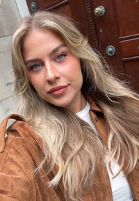 Brun semsket skinnjakke med krage, hvit topp under, langt bølgete blond hår, klare blå øyne, og en tre dør i bakgrunnen.