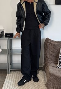 Schwarze Leder-Bomberjacke mit Lammfellfutter, schwarzes eng anliegendes T-Shirt, lockere schwarze Hose und schwarze Sneakers mit weißen Streifen.