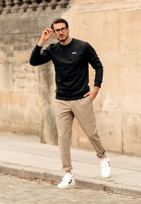 Sweatshirt preto de gola redonda, calças bejes ajustadas e ténis brancos com detalhes pretos. O modelo ajusta os óculos contra uma parede de pedra texturizada.
