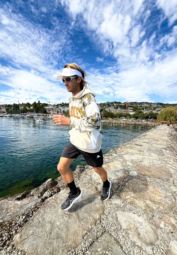 Homme portant des lunettes de soleil et une visière en train de faire du jogging sur un chemin de pierre au bord d'un plan d'eau avec des bateaux et des bâtiments en arrière-plan sous un ciel bleu.