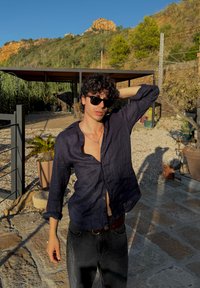 Camisa de lino azul marino con diseño de botón, mangas largas y cuello abierto. Combinada con jeans de mezclilla oscuros, situada en un área exterior pavimentada de piedra.