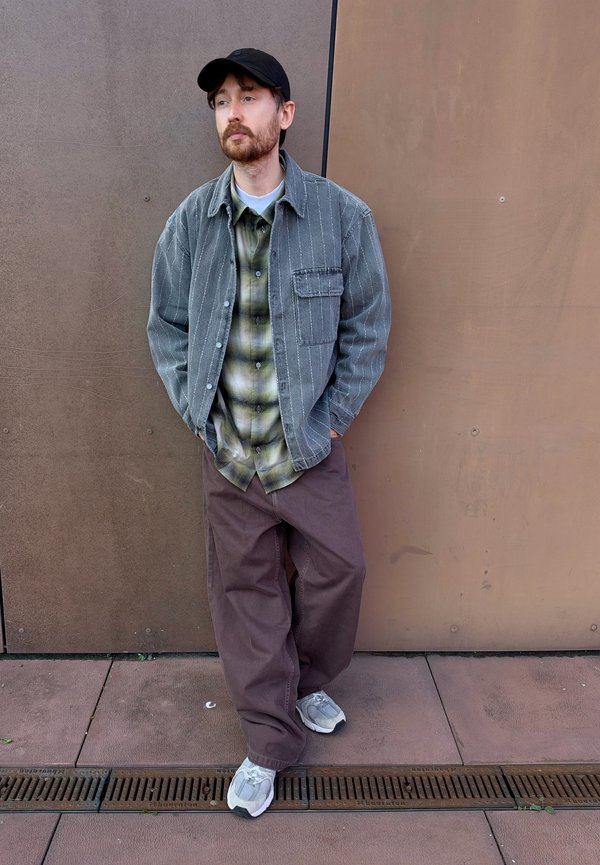 Un homme barbu portant une casquette noire, une veste et une chemise superposées, un pantalon ample et des baskets s’appuie contre un mur brun, les mains dans les poches.