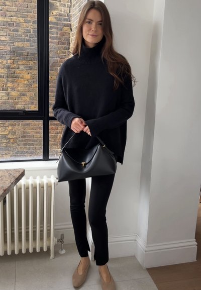 Mujer con suéter de cuello alto negro y pantalones, sosteniendo un bolso negro, parada en el interior junto a una ventana con una pared de ladrillos en el exterior.