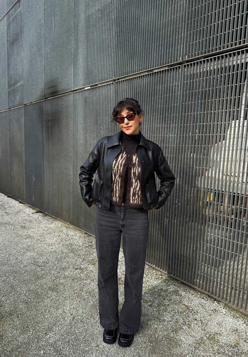 Black leather jacket over a patterned brown and black sweater, paired with gray wide-leg jeans and chunky black shoes. Sunglasses complete the look.