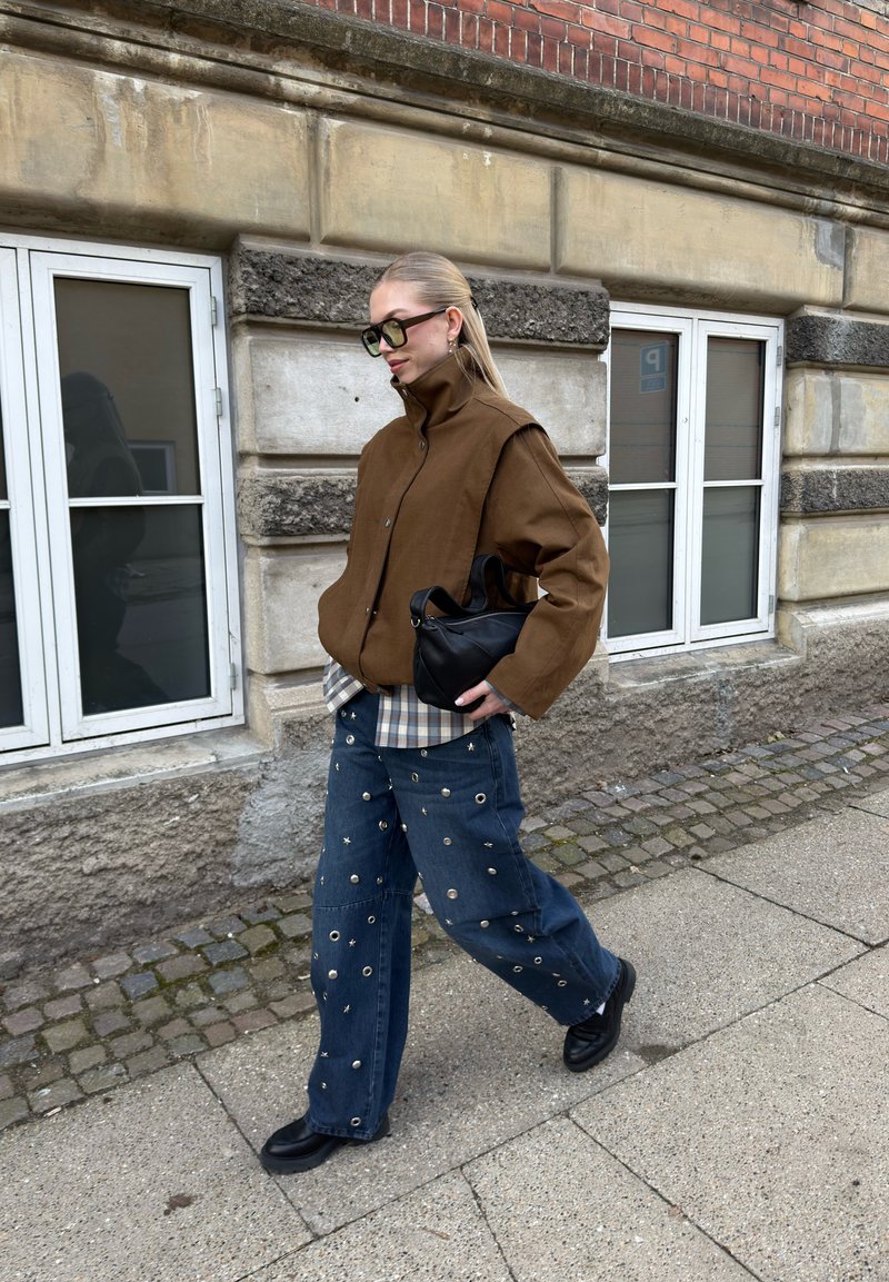 Femme portant une veste marron, une chemise à carreaux, un jean bleu avec œillets métalliques, des chaussures noires, portant un sac à main noir, marchant sur le trottoir de la ville.