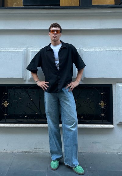 Camisa negra de manga corta sobre una camiseta blanca, jeans de pierna ancha de color azul claro, zapatillas verdes y gafas de sol con tintado naranja.
