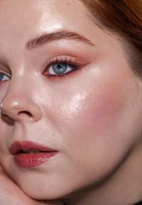 Nærbilde av et ansikt med glatt, opplyst hud, lys ferskenfarge rouge, dype blå øyne og myk rosa leppestift; subtile detaljer i øyemakeup.