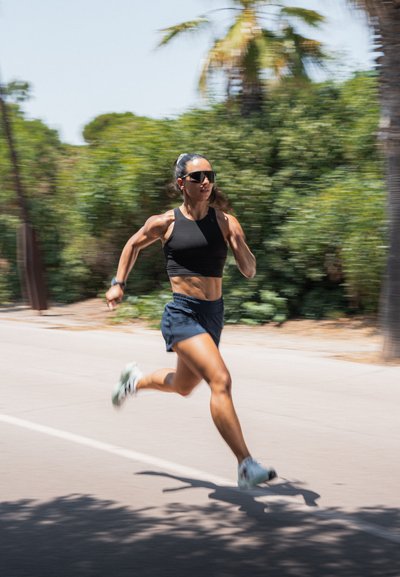 Atleta con un top corto negro y shorts azul marino corriendo por una carretera. Presenta unas gafas de sol elegantes y una pose dinámica, con vegetación de fondo.