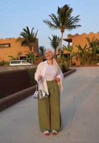 Γυναίκα με λευκό πουκάμισο, ελαιόχρωμα παντελόνια σε φαρδιά γραμμή και ασημένια τσάντα. Στο παρασκήνιο φαίνονται φοίνικες και κτήρια με υφή. Φωτεινός εξωτερικός χώρος.