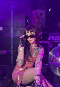Uma pessoa vestida com um vestido rosa brilhante e meias de rede, com tatuagens coloridas, segurando óculos de sol pretos, posou numa discoteca com uma bola de discoteca.