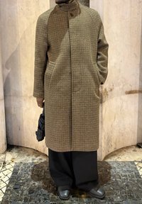 Personne portant un long manteau marron à carreaux, pantalons larges noirs, chaussures noires, tenant un parapluie noir plié, debout sur un pavé en pierre.