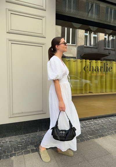 Vestido blanco texturizado con mangas abullonadas, cintura ceñida; bolso negro con herrajes plateados; zapatos gris de tipo slip-on; caminando junto a un escaparate.