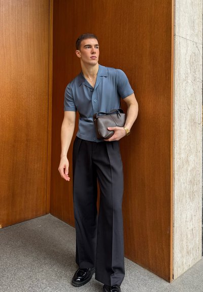 Camisa de manga corta azul claro, pantalones de pierna ancha negros y zapatos negros. Sostiene un pequeño bolso marrón oscuro. Textura suave, ajuste a medida.