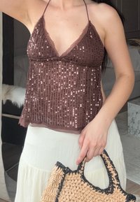 Donna che indossa un top a halter di paillettes marroni e una gonna bianca, mentre tiene una borsa di paglia intrecciata con rifiniture nere all'interno.