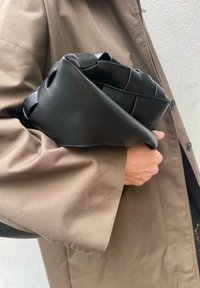 Black leather bag with a woven design, held under the sleeve of a brown coat. The bag boasts a smooth texture and contrasting stitching details.