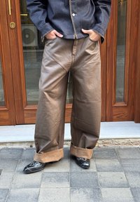 Pantalons large en cuir marron avec une taille haute, présentant des coutures visibles et des poignets retroussés, associés à des chaussures noires brodées.