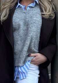 Pull gris côtelé porté sur une chemise à rayures bleu clair, assorti à un pantalon blanc, surmonté d'un manteau marron foncé. Main reposant dans la poche.