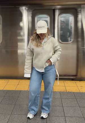Mujer con suéter beige, vaqueros azul claro y gorra blanca, de pie en la plataforma del metro con un tren detrás de ella.