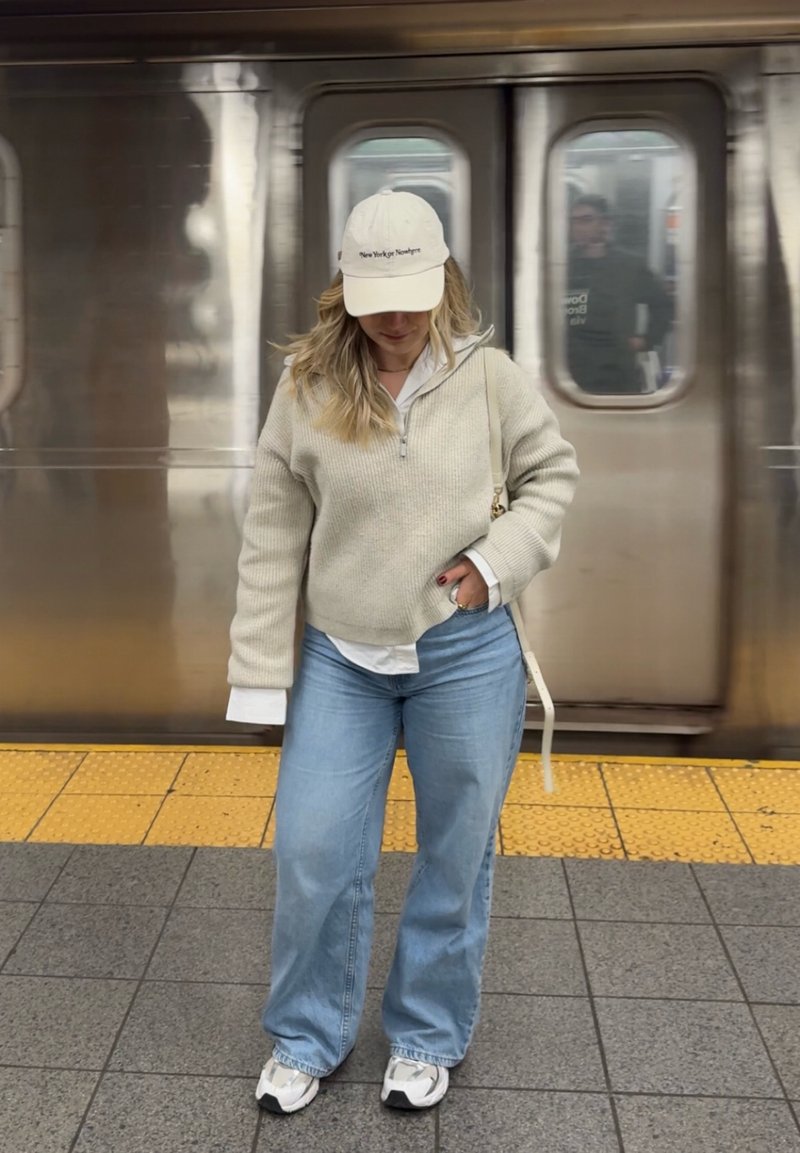 Femme portant un pull beige, un jean bleu clair et une casquette blanche, se tient sur le quai du métro avec un train derrière elle.