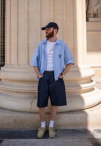 Hombre con barba que lleva una camisa de manga corta azul claro, camiseta blanca sin mangas, pantalones cortos de mezclilla oscura, zapatos beige y gorra azul marino, de pie junto a una gran columna de piedra.