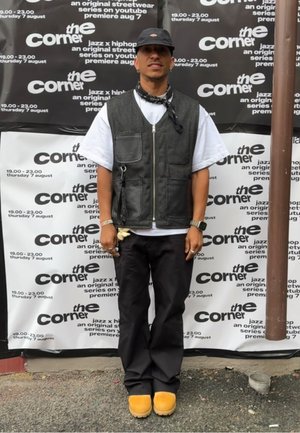 Black and gray sleeveless vest over a white shirt, black pants, and brown clogs. Accessories include a bandana and wristwatch. Background features event posters.