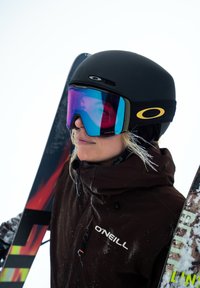 Casco da sci nero con visiera, occhiali da sci specchiati blu e viola, giacca marrone con logo, che tiene bastoncini da sci colorati, sfondo innevato.