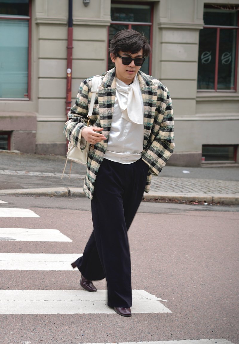 Personne portant des lunettes de soleil, un manteau à carreaux, un chemisier blanc et un pantalon noir traversant un passage piéton en ville avec un sac blanc à l’épaule.