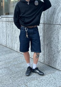 Schwarzer Kapuzenpullover, blaue Jeansshorts, schwarze Sneakers und weiße Socken. Auffälliges Tattoo am Bein und ein Schlüsselbund mit Schlüsseln an der Gürtelschlaufe.
