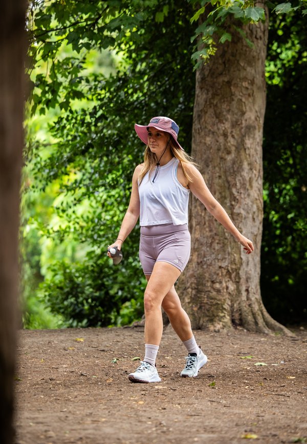 Mujer atlética con una camiseta sin mangas gris claro y pantalones cortos lavanda, usando un sombrero de ala ancha rosa, caminando con una botella de agua en la mano.